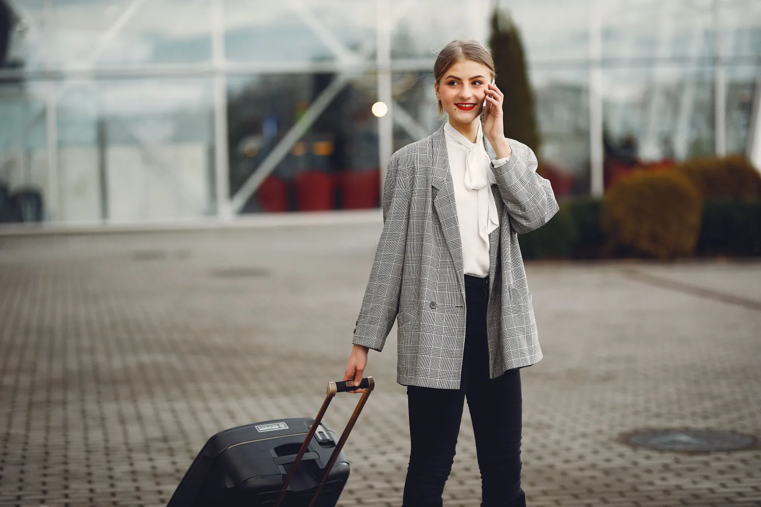 A traveller speaking on a phone while holding the handle of a suitcase outside a terminal.